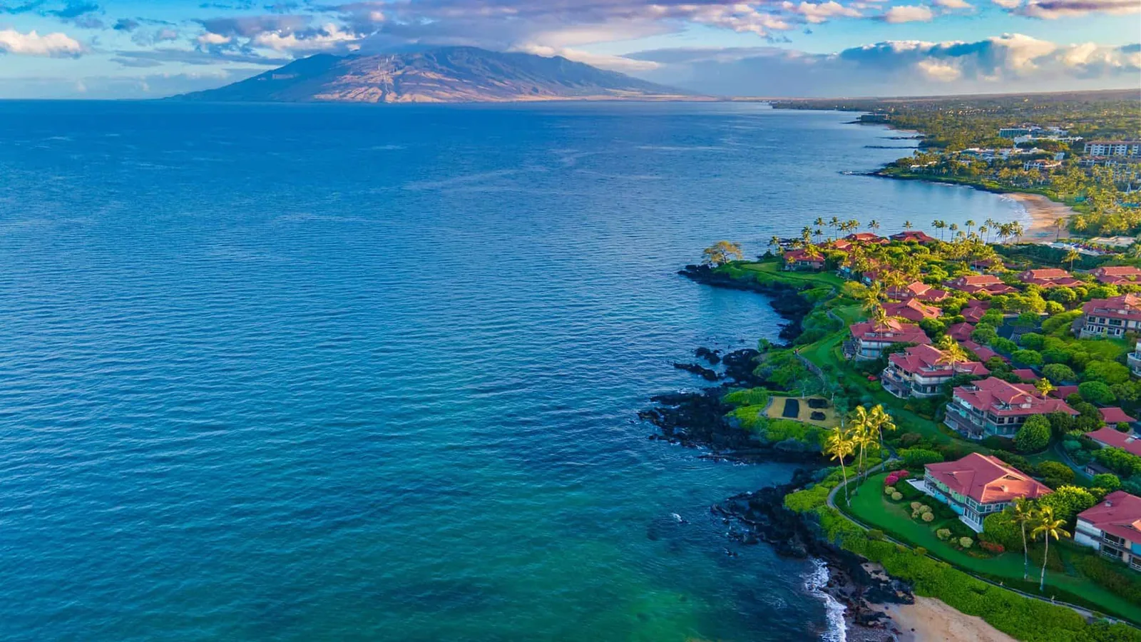 Aerial view of a South Maui coastline with turquoise water and oceanfront homes, the West Maui Mountains across the channel
